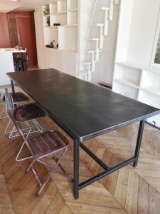 Table en métal patinée au milieu d’une salle à manger, avec chaises pliantes autour et étagères blanches en arrière-plan.