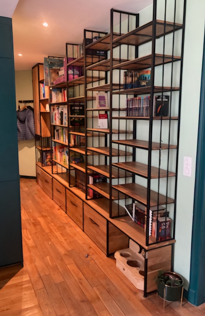 Étagères en bois massif et métal noir sur fond vert, rangées de livres et objets, rangement bas avec tiroirs, sol en parquet, atelier chaleureux Douceur Brute.